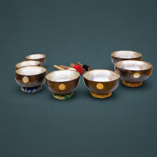 Seven chakra singing bowls with engraved sun and moon emblems resting on embroidered cushions with wooden mallets arranged behind them on a muted teal textured background. — for Carpe Diem Dream