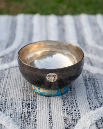 Hand-hammered Tibetan singing bowl with engraved sun and moon emblem resting on a blue embroidered cushion against a textured woven cloth background. — for Carpe Diem Dream