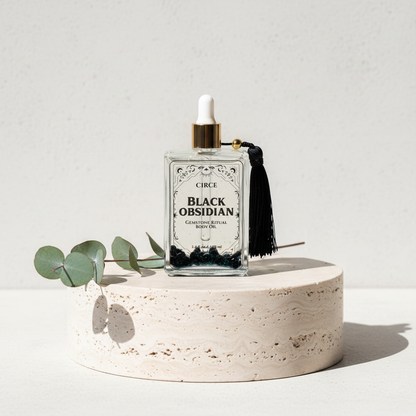 Black Obsidian Ritual Body Oil glass dropper bottle displayed on a travertine pedestal with eucalyptus leaves and soft botanical shadows. — for Carpe Diem Dream