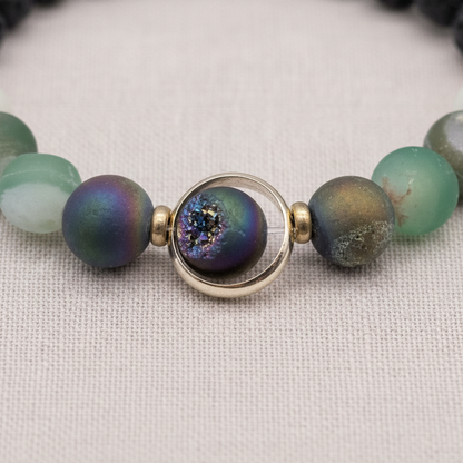 A close-up of a natural stone and lava bead diffuser bracelet featuring a round orbital focal bead with iridescent detailing and gold accents on a textured linen background. — for Carpe Diem Dream