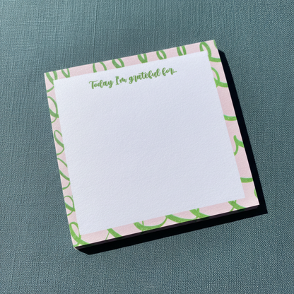 Close-up angled view of a pink and green gratitude sticky note pad with “Today I’m grateful for” text on a textured muted teal surface. — for Carpe Diem Dream