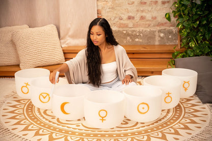 Woman seated on a patterned rug striking one of seven white frosted quartz crystal singing bowls with gold planetary symbols arranged in a circle — for Carpe Diem Dream
