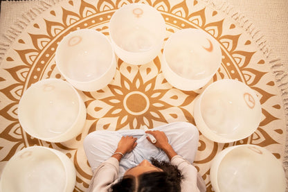 Top view of woman seated in meditation posture surrounded by seven white frosted quartz crystal singing bowls with gold planetary symbols arranged in a circle — for Carpe Diem Dream
