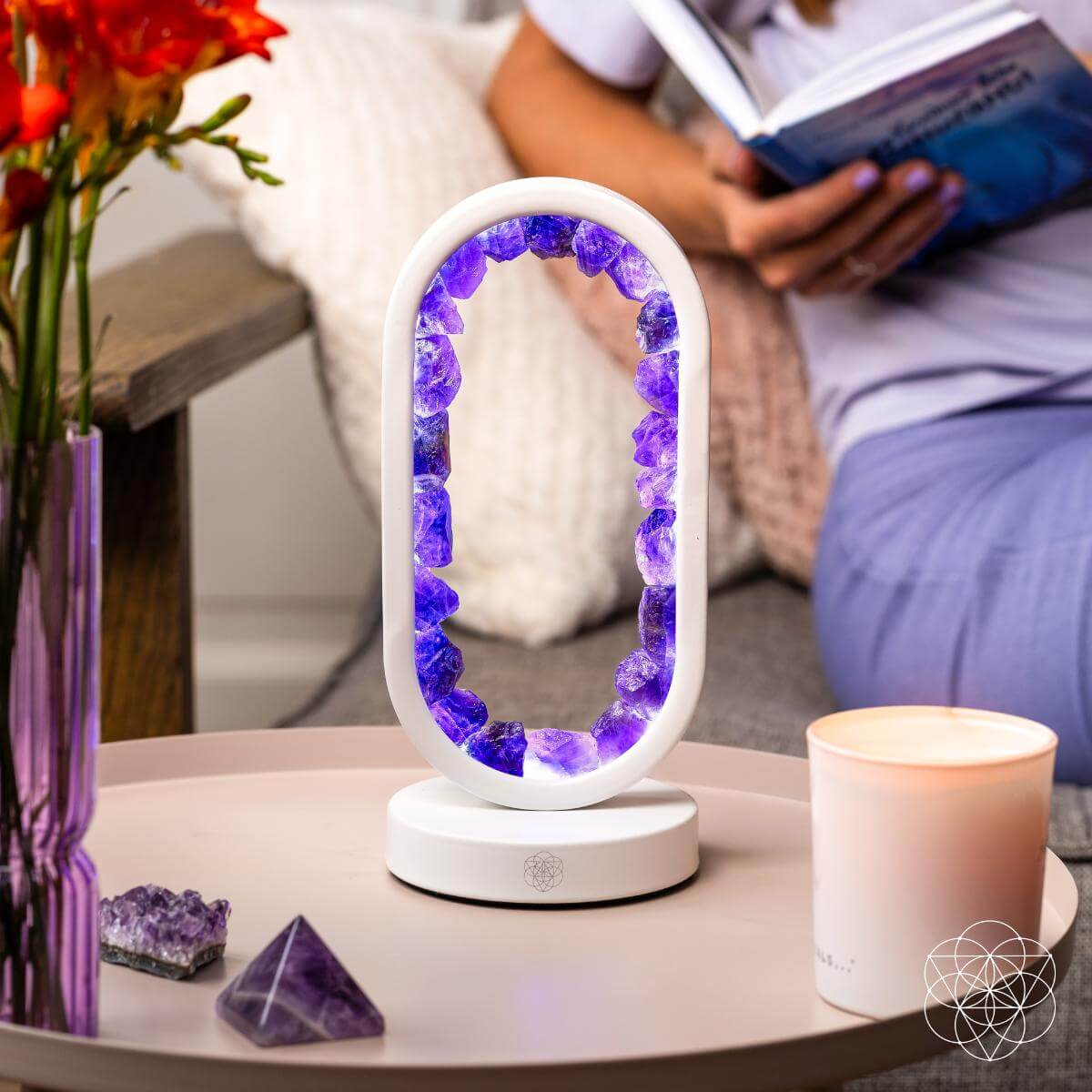 Decorative lamp with purple crystals on a table next to a vase of flowers and a candle, with a person reading a book in the background. Carpe Diem Dream