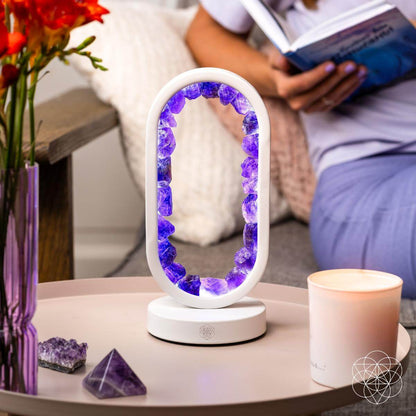 Decorative lamp with purple crystals on a table next to a vase of flowers and a candle, with a person reading a book in the background. Carpe Diem Dream