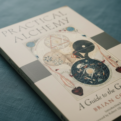 Close-up angled view of the paperback book Practical Alchemy featuring vintage alchemical artwork and serif typography resting on a muted teal linen-textured surface. — for Carpe Diem Dream
