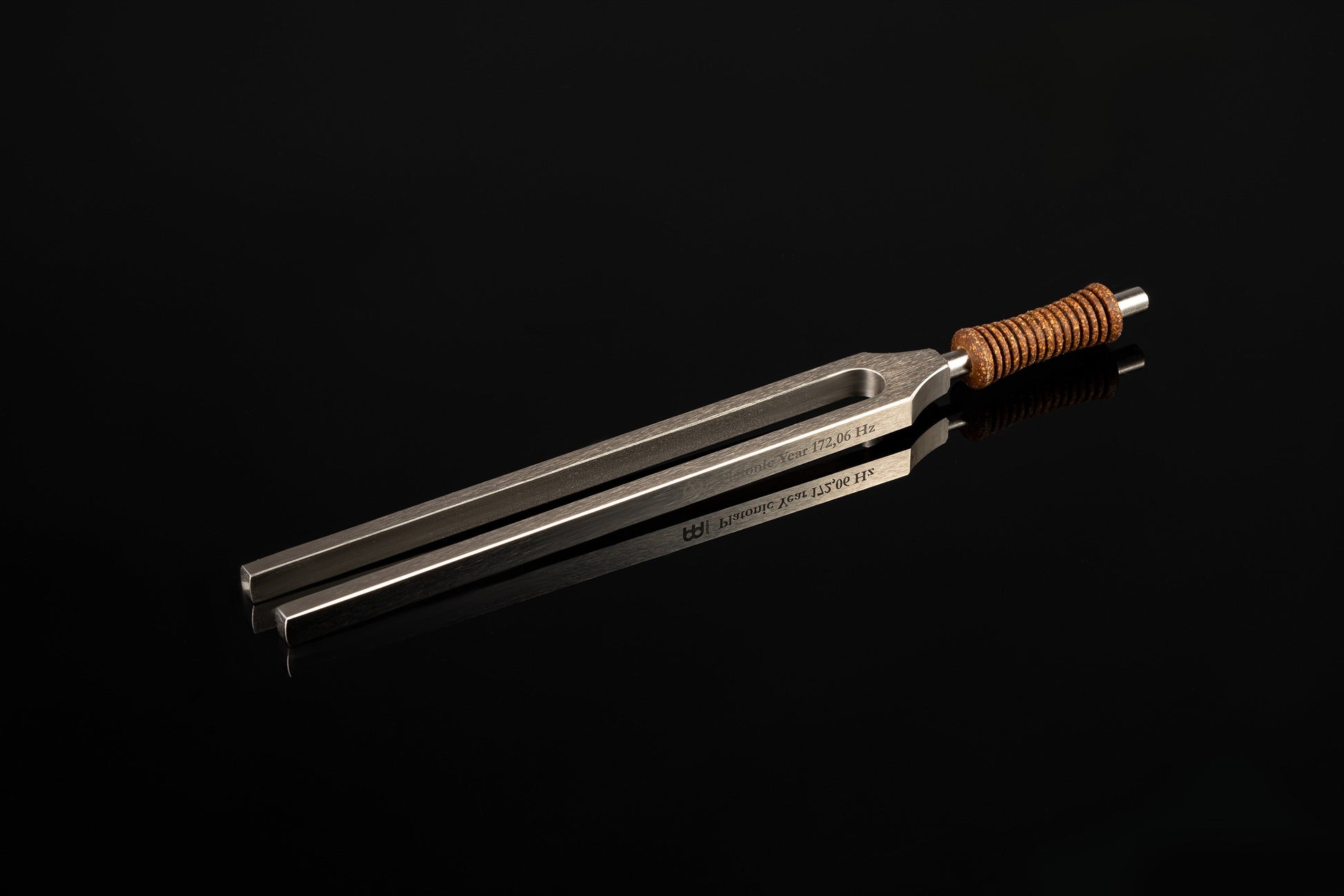 Weighted metal Platonic Year tuning fork engraved 172.06 Hz with cork handle resting on a glossy black reflective surface. — for Carpe Diem Dream