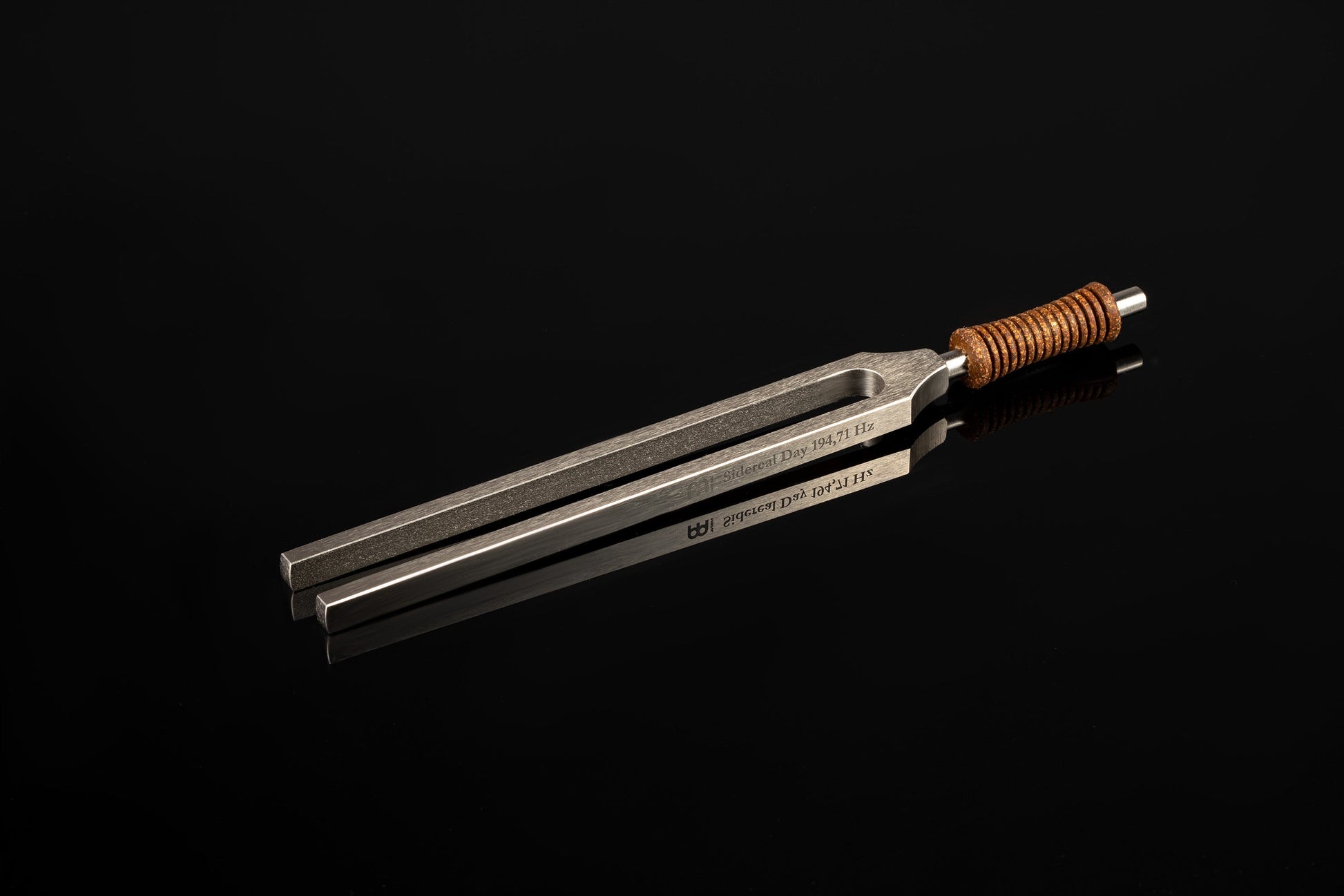 Weighted metal Sidereal Day tuning fork engraved 194.71 Hz with cork handle resting on a glossy black reflective surface. — for Carpe Diem Dream