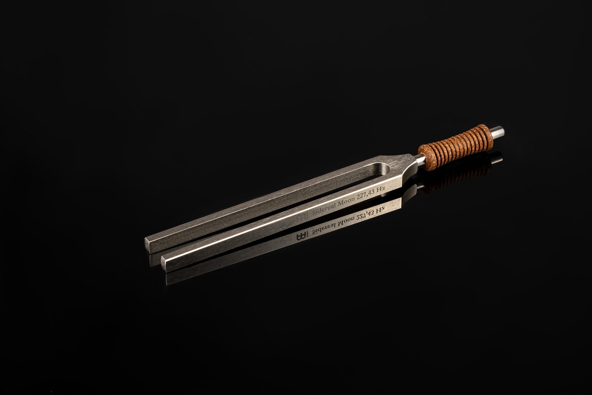 Weighted metal Sidereal Moon tuning fork engraved 227.43 Hz with cork handle resting on a glossy black reflective surface. — for Carpe Diem Dream