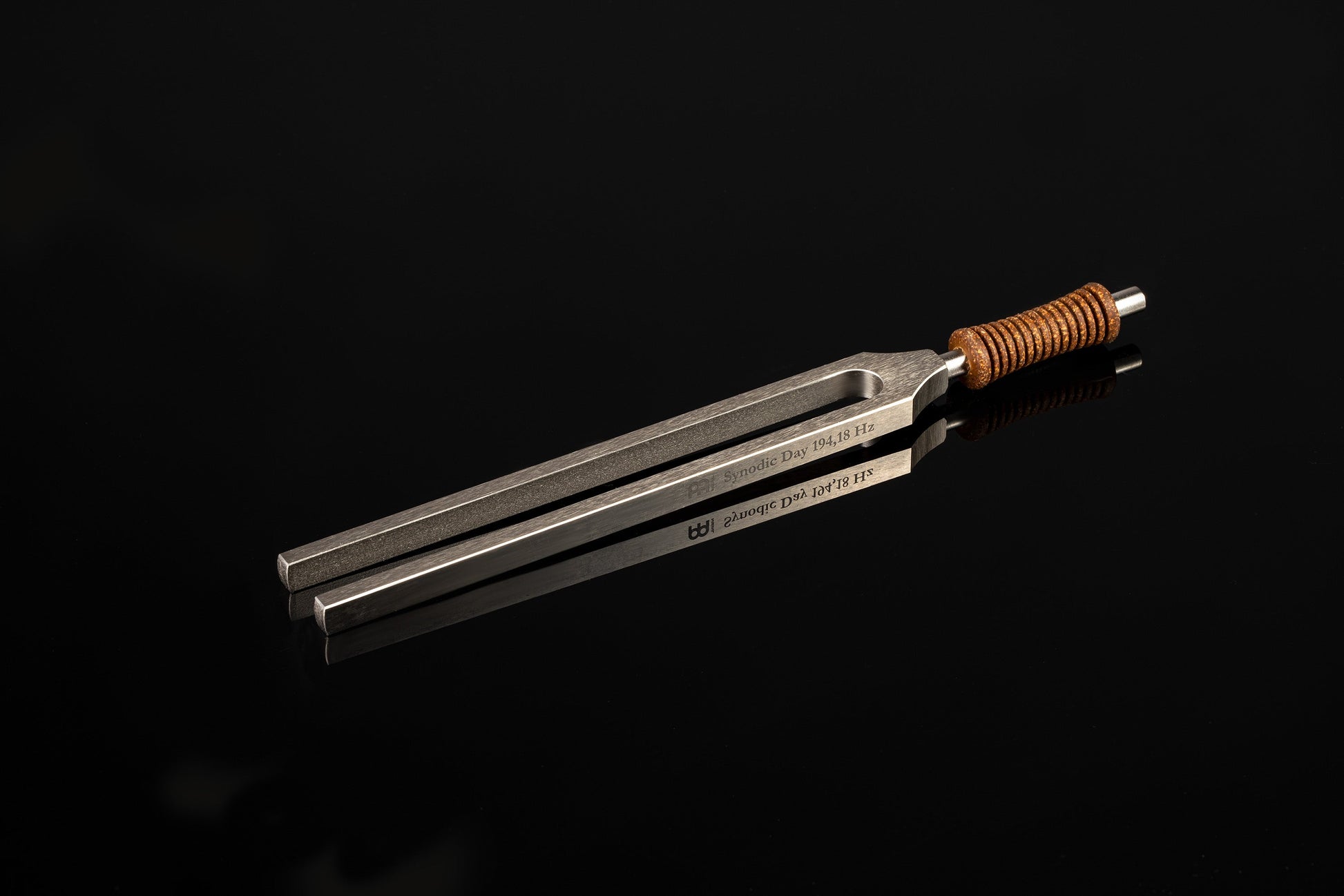 Weighted metal Synodic Day tuning fork engraved 194.18 Hz with cork handle resting on a glossy black reflective surface. — for Carpe Diem Dream
