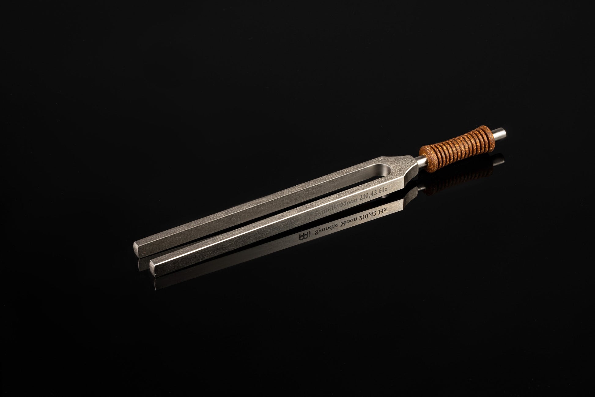 Weighted metal Synodic Moon tuning fork engraved 210.42 Hz with cork handle resting on a glossy black reflective surface. — for Carpe Diem Dream