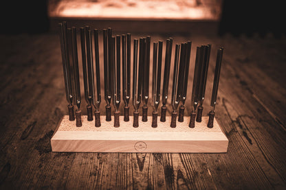 Sixteen weighted metal planetary tuning forks standing upright in a beech wood holder on a wooden table under warm lighting. — for Carpe Diem Dream