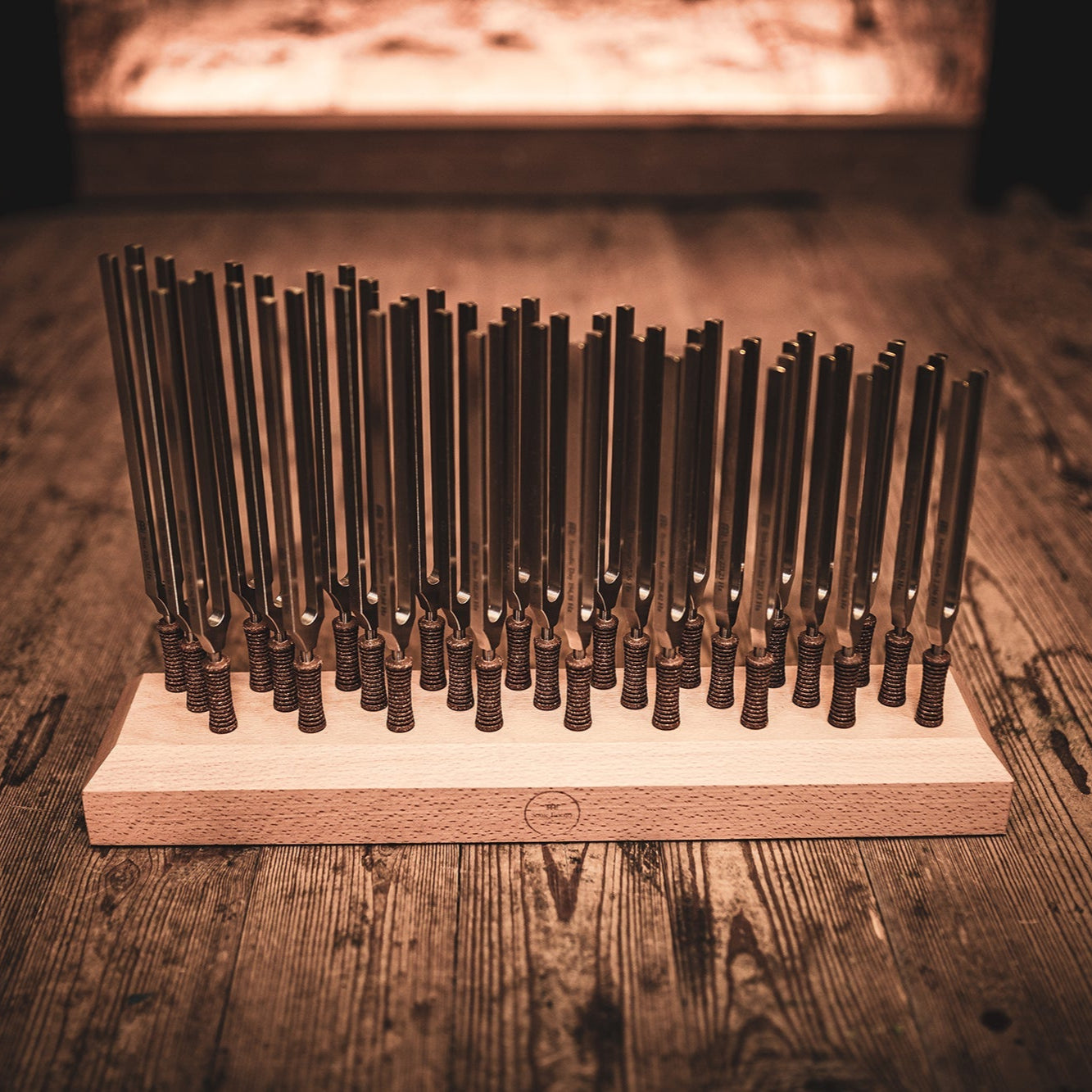 Twenty-seven weighted planetary therapy tuning forks standing upright in a wooden holder on a rustic wood surface with warm ambient lighting. — for Carpe Diem Dream