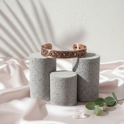 A pure copper floral cuff bracelet displayed on stone pedestals with soft botanical shadows, engraved leaf detailing, and neutral blush fabric styling. — for Carpe Diem Dream
