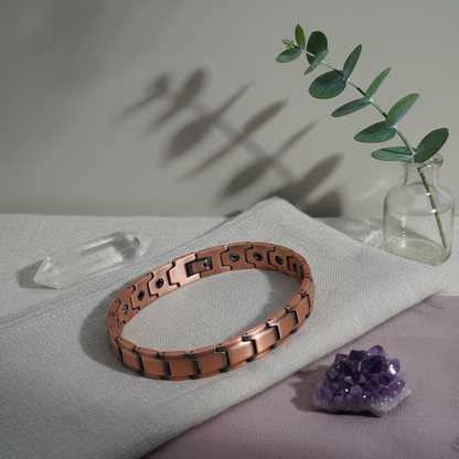 A pure copper magnetic bracelet resting on folded linen with a glass vase, green botanical stem, and crystals in soft natural light. — for Carpe Diem Dream