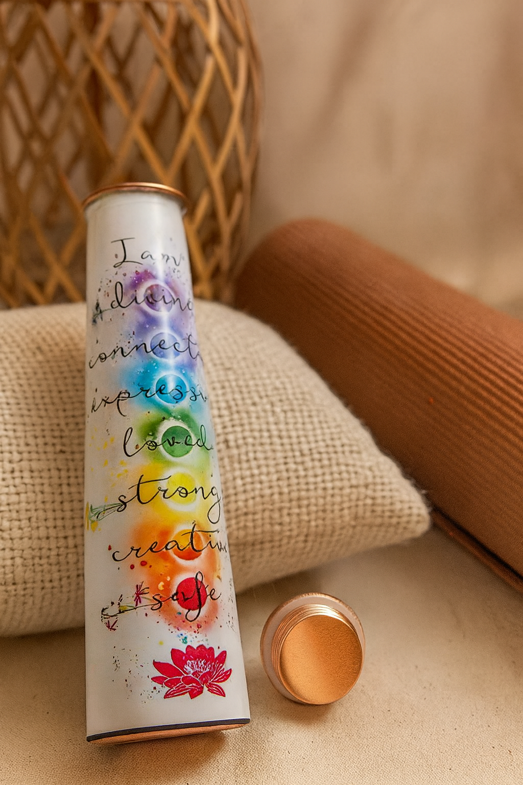 Pure copper water bottle with printed “I Am” chakra affirmations resting on cozy yoga cushions with its matching copper lid beside it in a warm neutral setting. — for Carpe Diem Dream