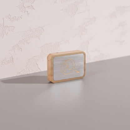 A Qi-Me EMF protection device with a brushed metal face and wood frame displayed on a clean neutral surface against a softly textured wall with gentle natural lighting. — for Carpe Diem Dream