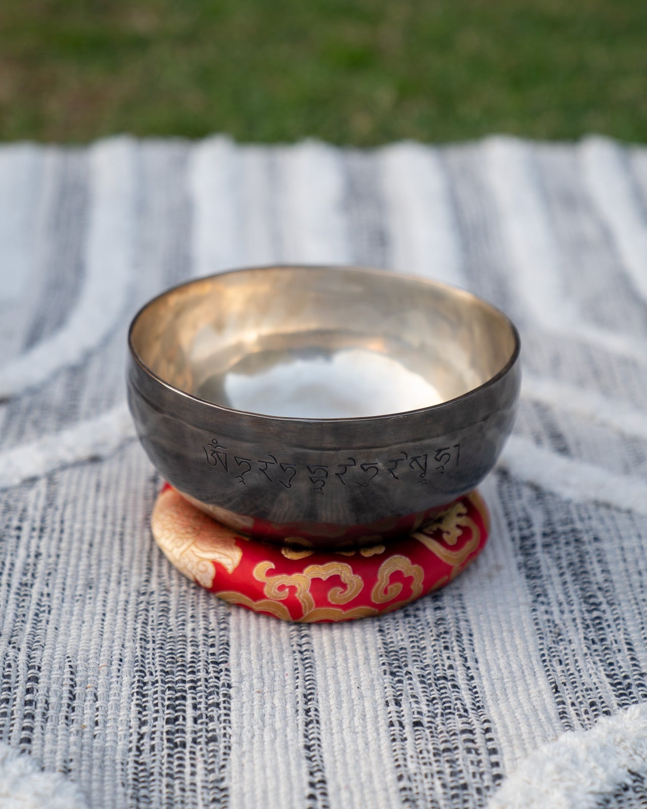 Back view of a Tibetan Root Chakra New Moon singing bowl with engraved mantra resting on a red patterned cushion on a textured woven blanket background. — for Carpe Diem Dream