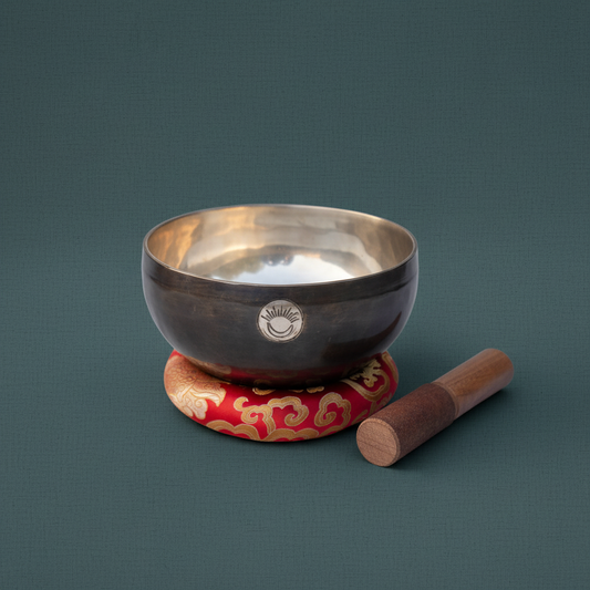 Root Chakra New Moon singing bowl with engraved sun and moon symbol resting on a red patterned cushion with wooden striker beside it on a muted teal background. — for Carpe Diem Dream