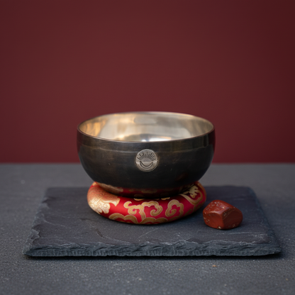Root Chakra New Moon singing bowl with engraved sun and moon symbol resting on a red patterned cushion displayed on a slate platform with a deep red studio background. — for Carpe Diem Dream