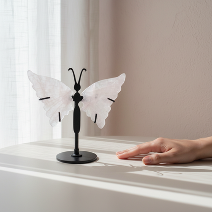 Rose quartz crystal butterfly wings on a black stand displayed next to a hand for size reference on a softly lit windowsill. — for Carpe Diem Dream