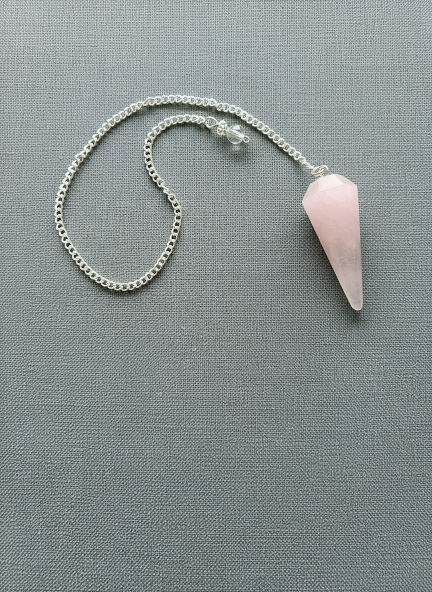 Rose Quartz crystal pendulum with silver chain displayed on a textured pale gray surface, radiating soft pink tones and gentle light – for Carpe Diem Dream.