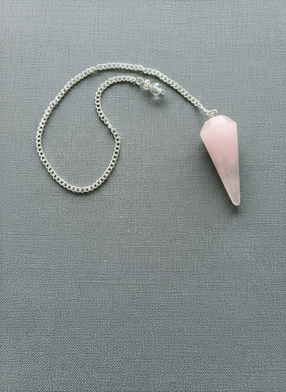 Rose Quartz crystal pendulum with silver chain displayed on a textured pale gray surface, radiating soft pink tones and gentle light – for Carpe Diem Dream.