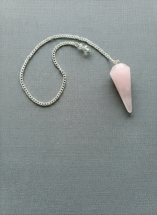 Rose Quartz crystal pendulum with silver chain displayed on a textured pale gray surface, radiating soft pink tones and gentle light – for Carpe Diem Dream.