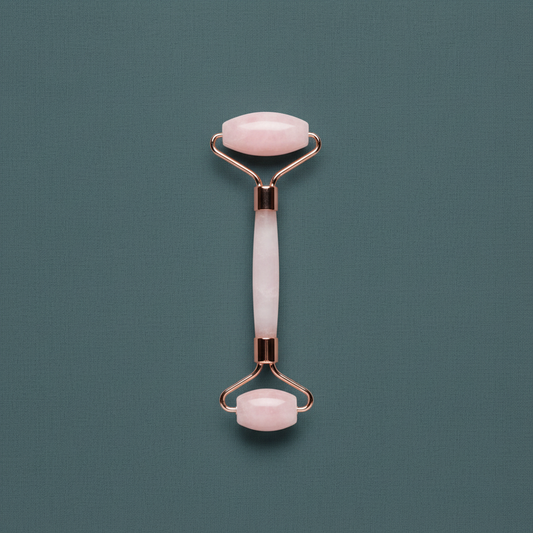 A dual-ended rose quartz facial roller with polished crystal rollers and rose gold frame displayed upright on a muted sage textured background. — for Carpe Diem Dream