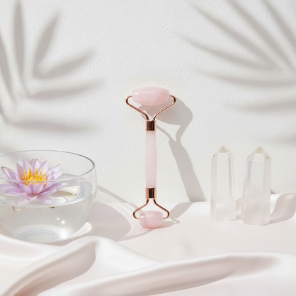 A rose quartz facial roller standing upright beside clear quartz crystals and a floating lotus flower bowl on a soft white botanical spa setting. — for Carpe Diem Dream
