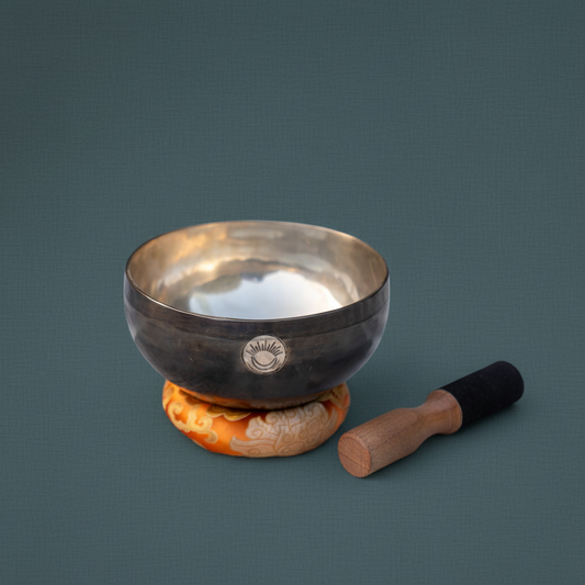 Sacral Chakra New Moon singing bowl with engraved sun and moon symbol resting on an orange cushion beside a wooden striker on a muted teal background. — for Carpe Diem Dream