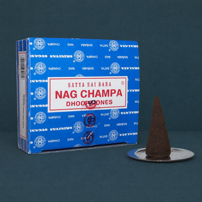 Satya Nag Champa dhoop cones box with one brown cone on a small metal disc burner against a dark teal textured linen background. — for Carpe Diem Dream