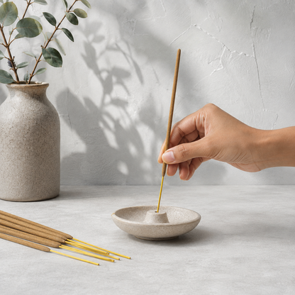 Hand placing an unlit Satya Nag Champa incense stick into a ceramic holder on a cool pale-stone surface. — for Carpe Diem Dream.