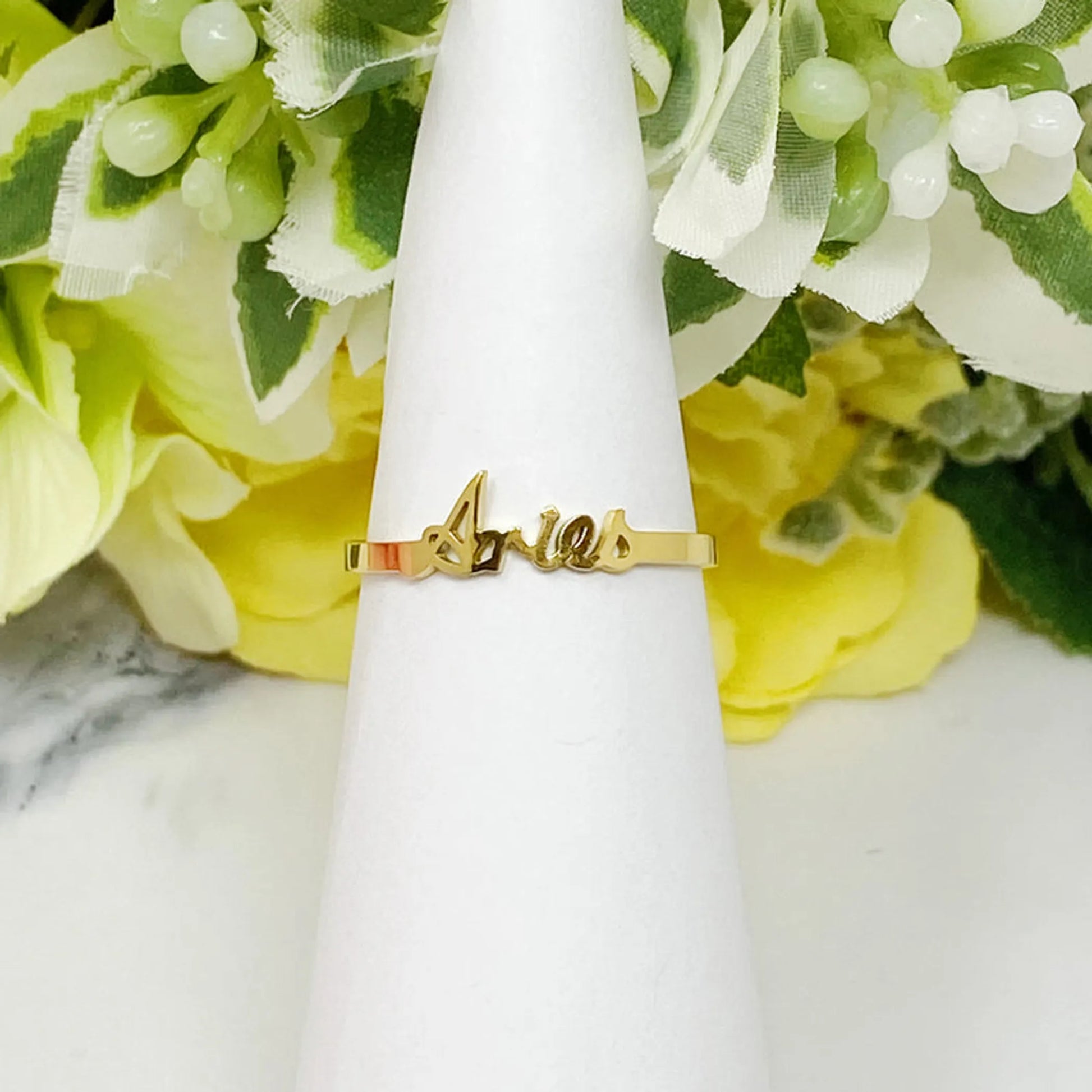 18K gold Scripted Zodiac Ring featuring delicate cursive lettering of zodiac Aries, displayed on white botanical background for Carpe Diem Dream