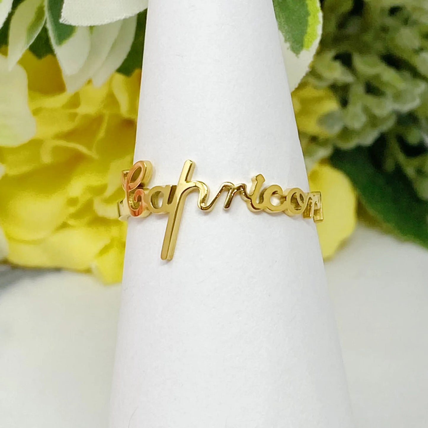18K gold Scripted Zodiac Ring featuring delicate cursive lettering of zodiac Capricorn, displayed on white botanical background for Carpe Diem Dream