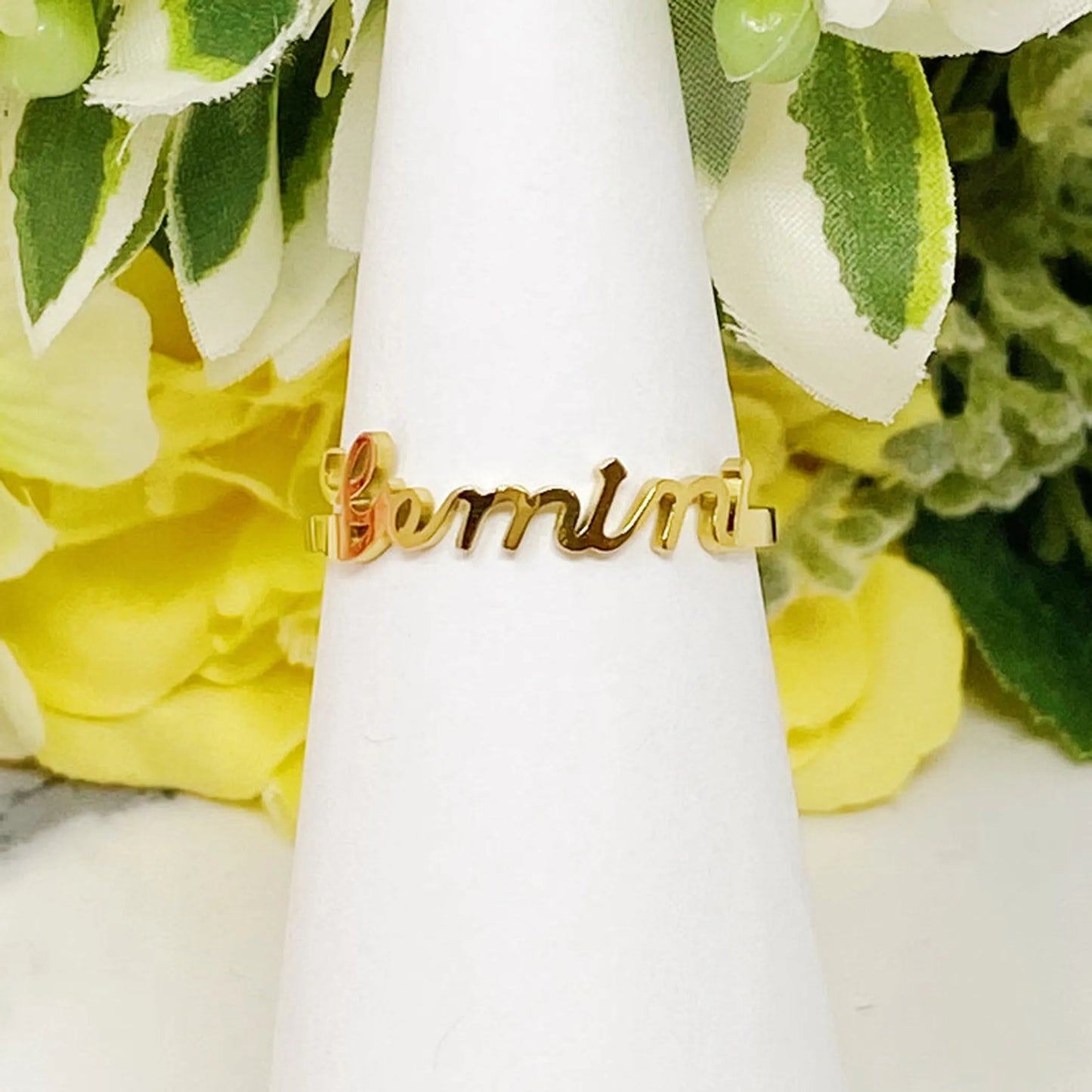 18K gold Scripted Zodiac Ring featuring delicate cursive lettering of zodiac Gemini, displayed on white botanical background for Carpe Diem Dream