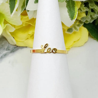 18K gold Scripted Zodiac Ring featuring delicate cursive lettering of zodiac Leo, displayed on white botanical background for Carpe Diem Dream