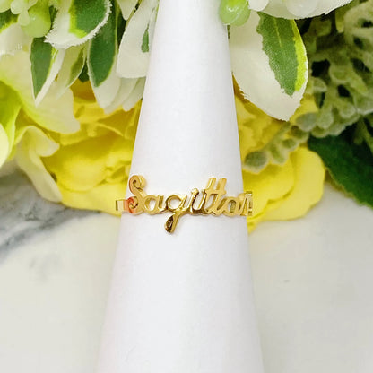 18K gold Scripted Zodiac Ring featuring delicate cursive lettering of zodiac Sagittarius, displayed on white botanical background for Carpe Diem Dream
