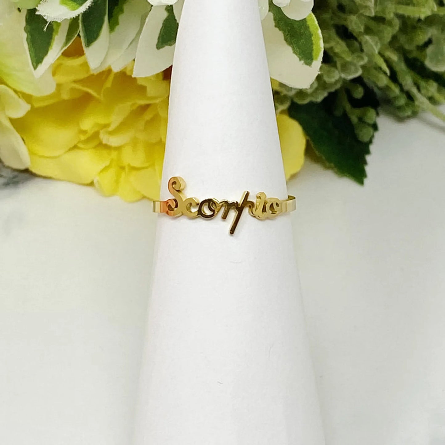 18K gold Scripted Zodiac Ring featuring delicate cursive lettering of zodiac Scorpio, displayed on white botanical background for Carpe Diem Dream