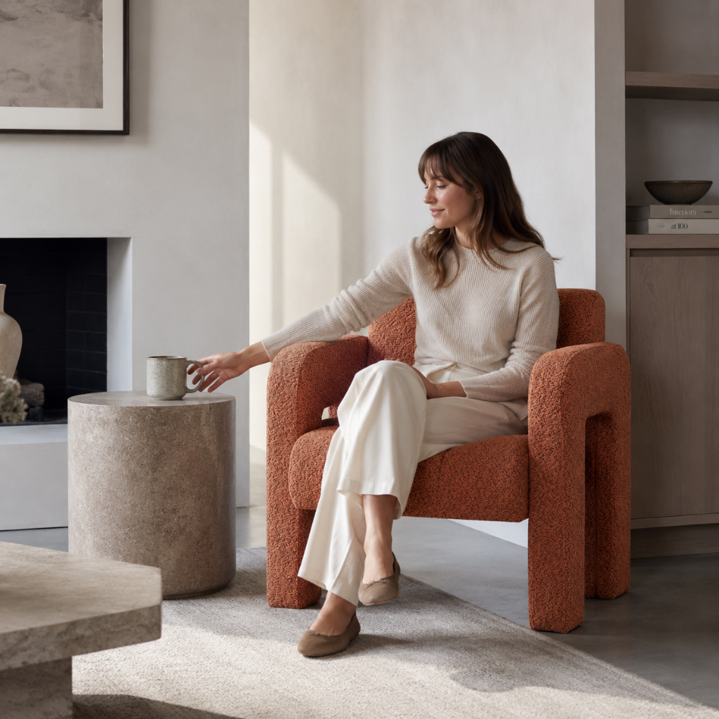 Woman seated in the orange sculpted bouclé upholstered accent chair while reaching for a ceramic mug in a refined living room with light greige walls, soft stone surfaces, warm white textiles, and cool natural light. — for Carpe Diem Dream
