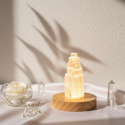 Selenite cleansing crystal lamp glowing on a round wooden base with clear quartz points and white flower in a soft botanical studio setting. — for Carpe Diem Dream
