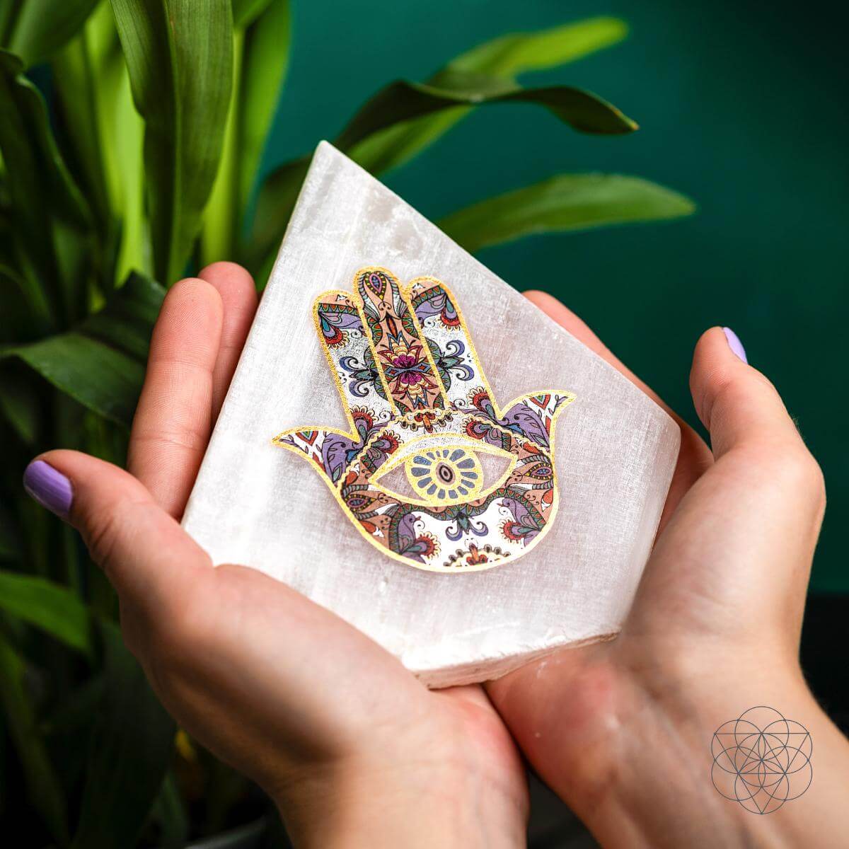Selenite crystal lamp featuring a detailed Hamsa Hand design, held gently in both hands against a lush green background — symbolizing protection, mindfulness, and calm energy, styled by Carpe Diem Dream.