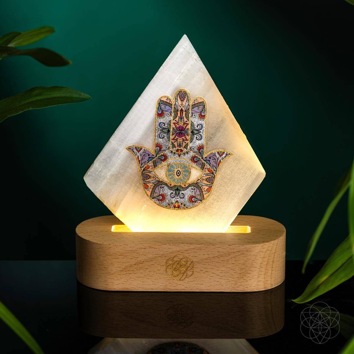 Selenite Hamsa Mood Lamp glowing softly against a dark green background, resting on a wooden base. The luminous Hamsa design radiates protection, peace, and mindful serenity