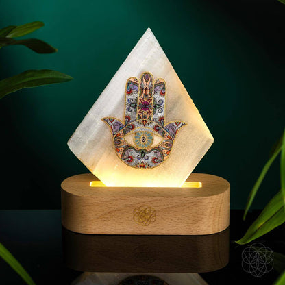 Selenite Hamsa Mood Lamp glowing softly against a dark green background, resting on a wooden base. The luminous Hamsa design radiates protection, peace, and mindful serenity
