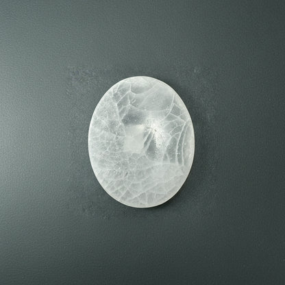 An oval selenite slice with a soft translucent texture displayed upright on a muted dark textured background. — for Carpe Diem Dream