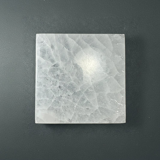 A square selenite slice with a soft translucent texture displayed upright on a muted dark textured background. — for Carpe Diem Dream