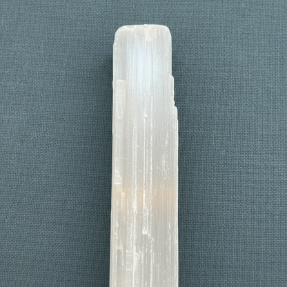 A close-up view of a rough selenite stick showing natural striations and translucent texture against a muted teal fabric background. — for Carpe Diem Dream