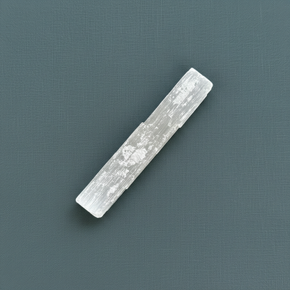 A rough selenite stick displayed diagonally on a muted teal textured background. — for Carpe Diem Dream
