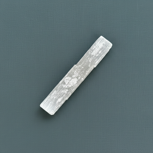A rough selenite stick displayed diagonally on a muted teal textured background. — for Carpe Diem Dream
