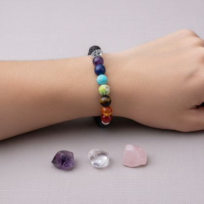 A person wearing a black lava stone chakra bracelet with multicolored beads and a silver Tree of Life charm above three small crystals on a neutral background. — for Carpe Diem Dream